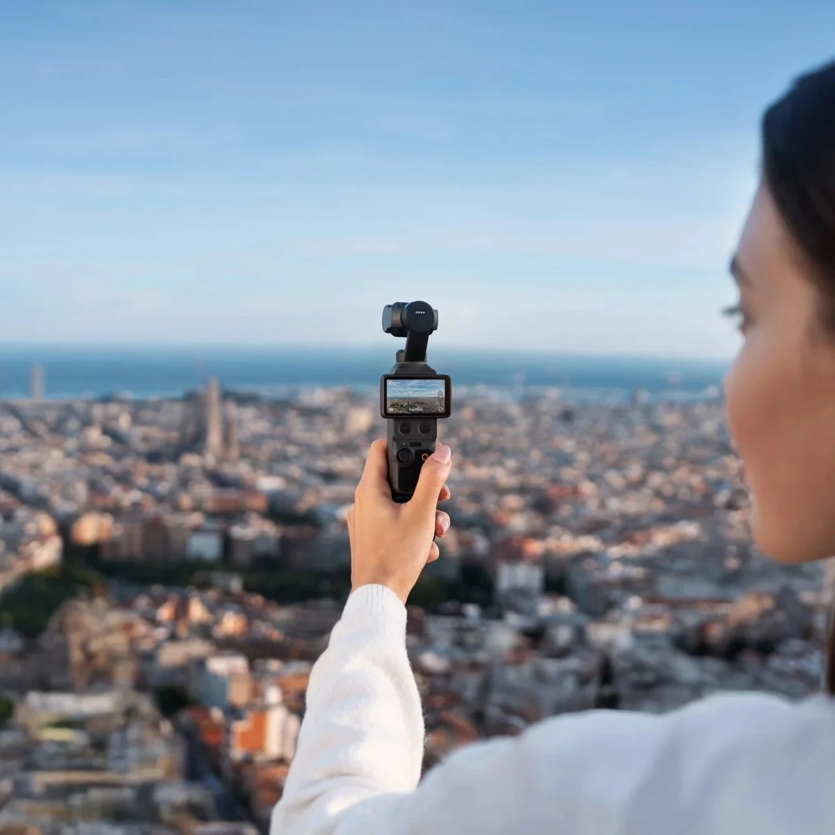Женская рука держит камеру с трехосевым стабилизатором DJI Osmo Pocket 4 с развернутым горизонтальным дисплеем на фоне городской панорамы